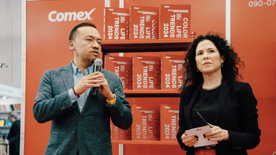 Hombre vestido de gris y mujer vestida de negro, en el fondo se ven unos libros de la marca Comex en color naranja.