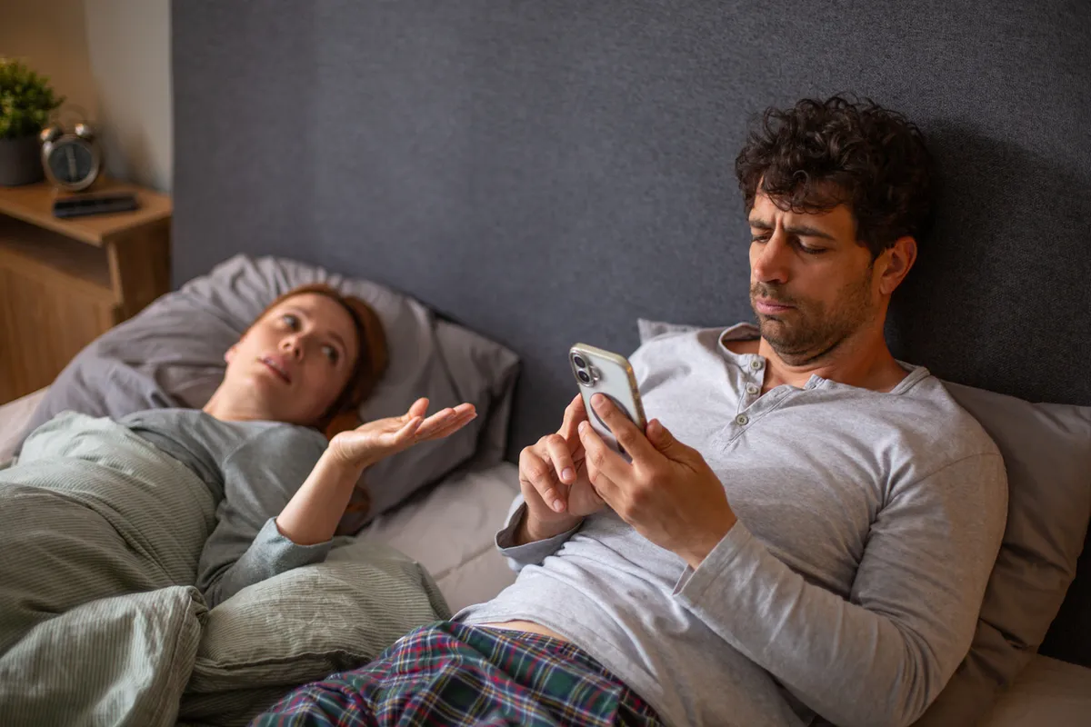 Man ignoring woman in bed while phubbing using smartphone