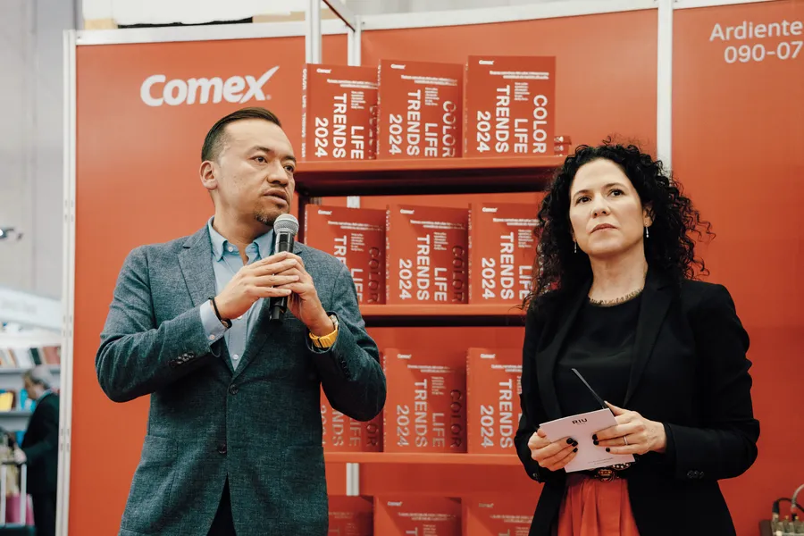 Hombre vestido de gris y mujer vestida de negro, en el fondo se ven unos libros de la marca Comex en color naranja.